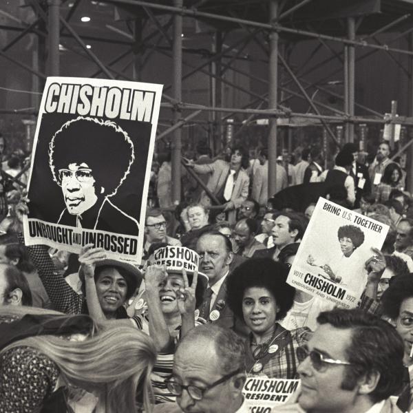 A black and white photograph of a crowd gathered in support of candidate Shirley Chisholm. Several people are smiling and holding up signs supporting her campaign for president.