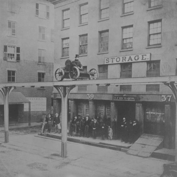 Black and white photo of a man riding in a cart on a elevated platform above a city street.