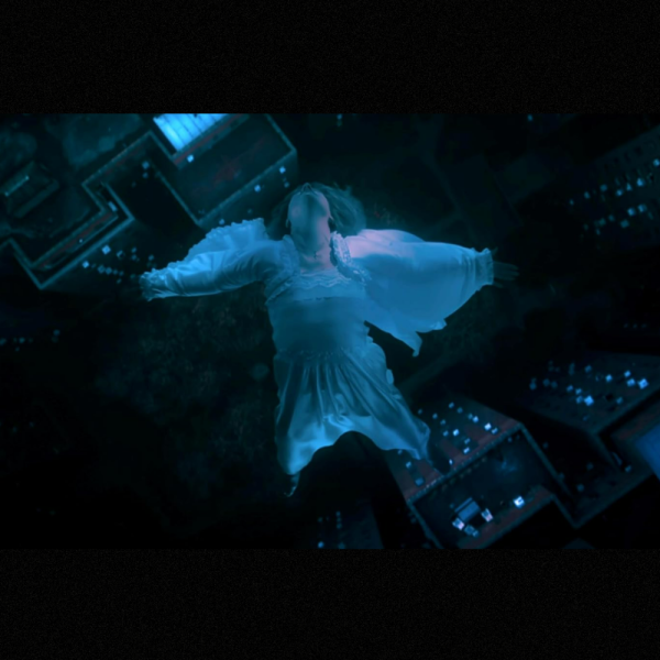 A woman in a flowing white dress appears to float or fall gracefully in a dimly lit urban nightscape, viewed from above.