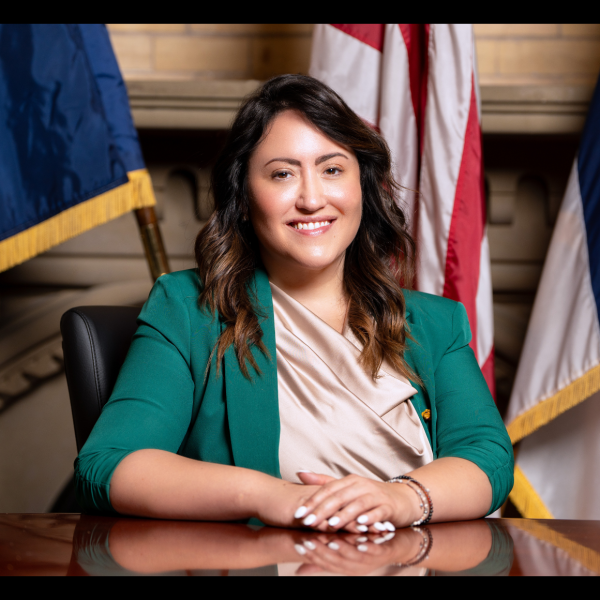 Woman in a green suit jacket sitting at a desk smiling in front of the American flag 