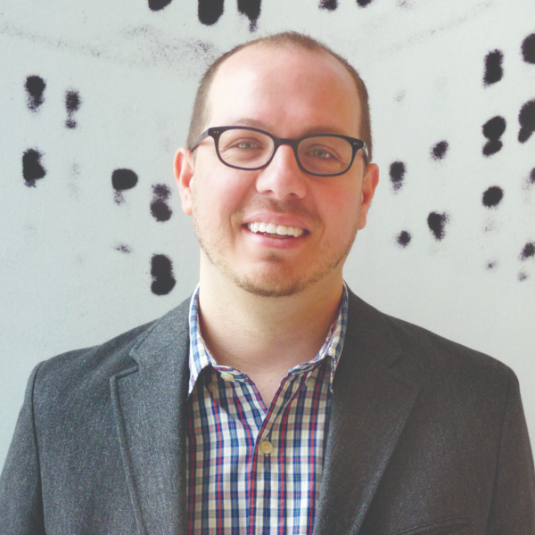 Man wearing glasses and a grey suit smiling in front of a black and white wall