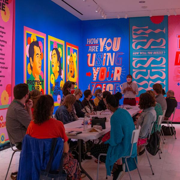 A group of educators sitting around a table listening to a facilitator against a backdrop of a brightly colored mural.