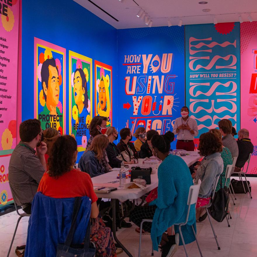 A group of educators sitting around a table listening to a facilitator against a backdrop of a brightly colored mural.