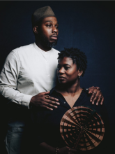 Tanika, a Black woman wearing a black shirt, sits holding a circular pleated fan. She looks to the left. Shakeem, a Black man wearing a white shirt and olive green hat stands with his hands on her shoulders, looking to the left. Behind them is a dark blue background.