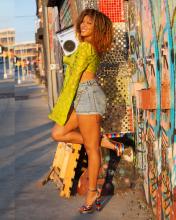 DJ Taela Naomi kicks her leg up in front of a mosaic wall. She wears a line green lace bell sleeved top, denim shorts, and rainbow heels. She holds a boom box on her shoulder and smiles at the camera.