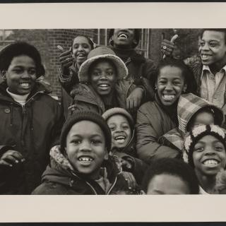 Black and white photograph of a group of Black-presenting children wearing winter clothes and smiling at the camera