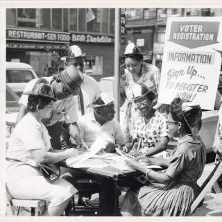 A black and white photograph of eight people sitting around a table on a city sidewalk. In the middle of the group is Shirley Chisholm, sitting under a sign saying, “Voter Registration Information.