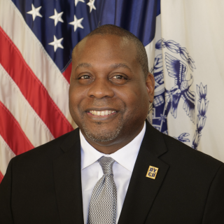 A man is wearing a suit and smiling in front of the American Flag