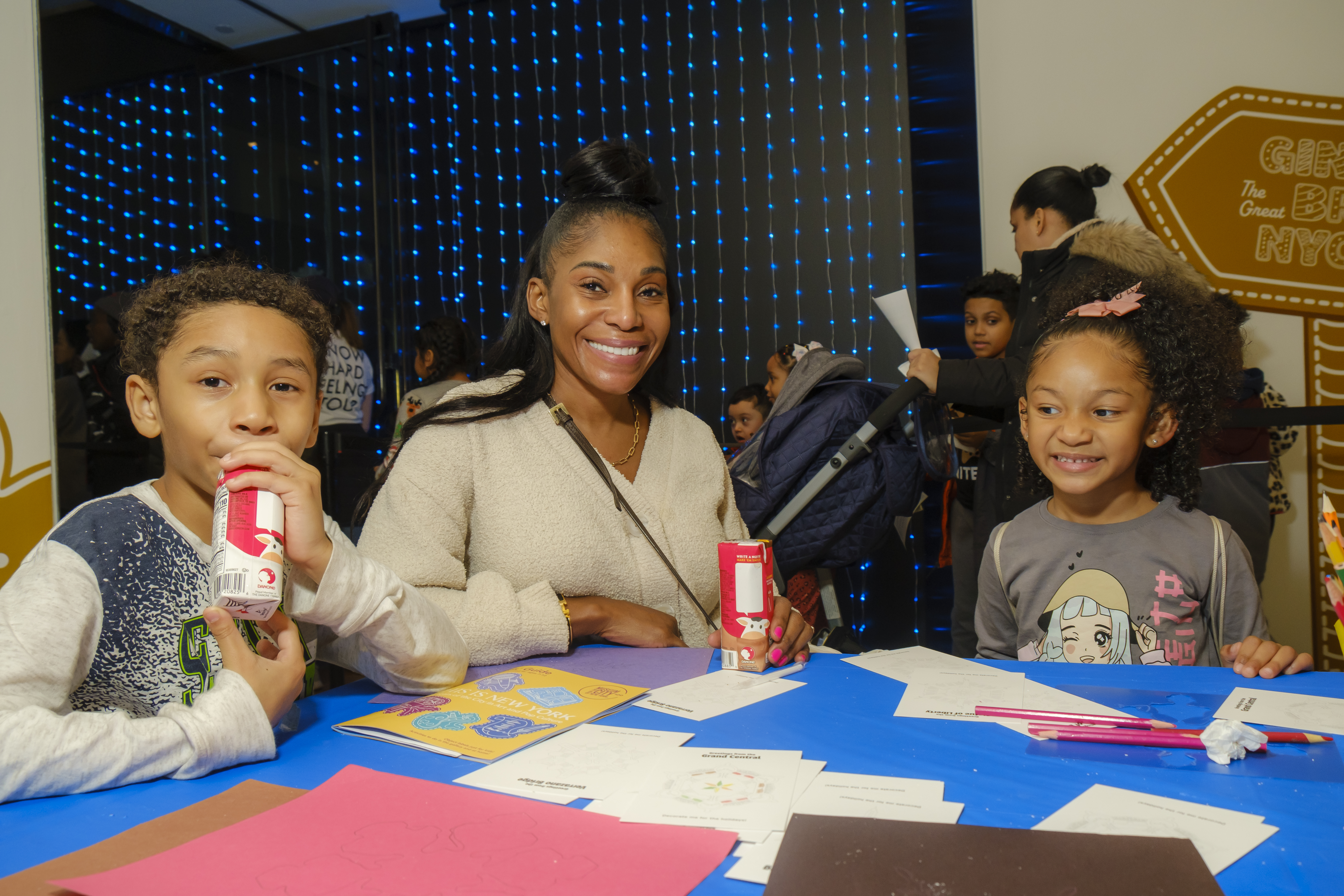 A woman and two young children smile as they make art at a table.